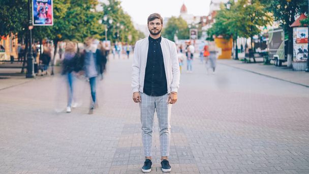 Portrait of young businessman in trendy garments standing in busy pedestrian street and looking at camera while crowds of people are whizzing around. - Photo, image