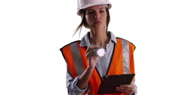 Vrouwelijke werknemer in veiligheidsuitrusting met tablet en zaklamp op effen wit kopieerruimte. Vrouwelijke ingenieur werkt schijnende zaklamp op camera op witte achtergrond. 4k - Video