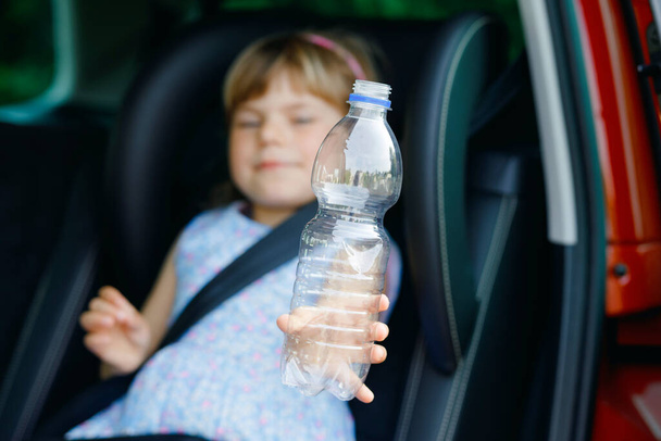 Entzückendes Kleinkind, das im Autositz sitzt, eine Flasche mit Wasser in der Hand hält und aus dem Fenster auf Natur und Verkehr blickt. Kleines Kind, das mit dem Auto reist. Kindersicherheit im Straßenverkehr. Familienausflug und Urlaub. - Foto, Bild