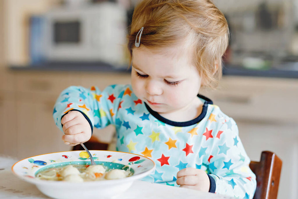 Очаровательная малышка ест с ложки овощного супа с лапшой. питание, ребенок, питание и концепция развития. Симпатичный малыш, дочь с ложкой сидит в кресле и учится есть самостоятельно - Фото, изображение