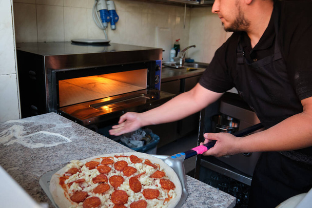 Latin man opening the oven to cook pepperoni pizza in a restaurant kitchen. Pizzeria. - Photo, Image