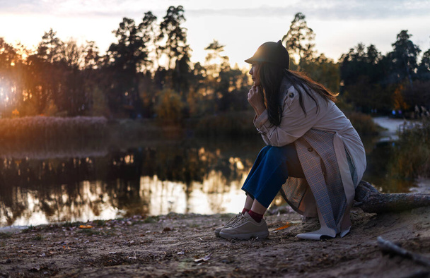 秋に森の中で目を閉じて大きな木の近くに座ってコートを着た若いアジアの女性. - 写真・画像