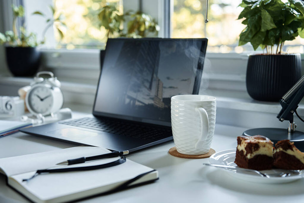 Miejsce pracy w biurze. Stół z laptopem, notatnikiem i gorącą kawą przy oknie w salonie. - Zdjęcie, obraz