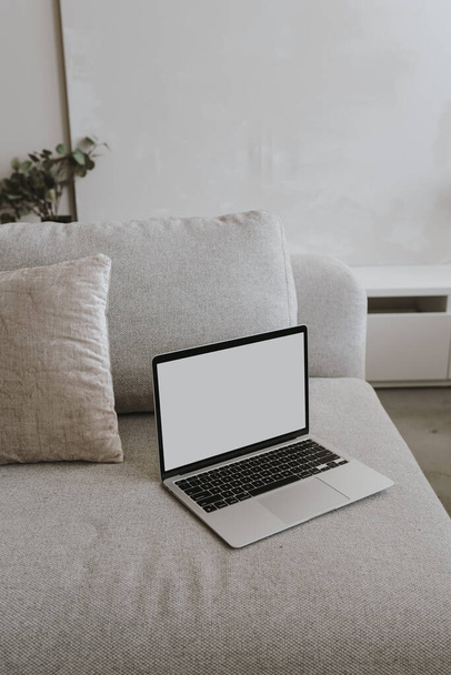Laptop-Computer mit leerem Kopierplatz-Bildschirm auf einem bequemen Sofa mit Kissen. Ästhetische Wohnzimmereinrichtung. Online-Shopping, Online-Shop, soziale Medien, Blog-Branding-Attrappe - Foto, Bild