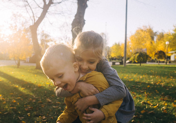 Брат і сестра в парку. Хлопчик і дівчинка тримаються за руки і бігають по дорозі до парку. Концепція щасливого дитинства та гетеросексуальних родичів. Сонячні промені і жовте листя
 - Фото, зображення