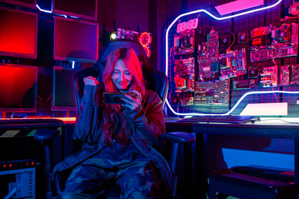 Winning. Happy Gamer playing video game online with smartphone with neon lights she raises hands to wins celebrating, confused Asian woman wearing gaming headphones feeling excited at home - Photo, image