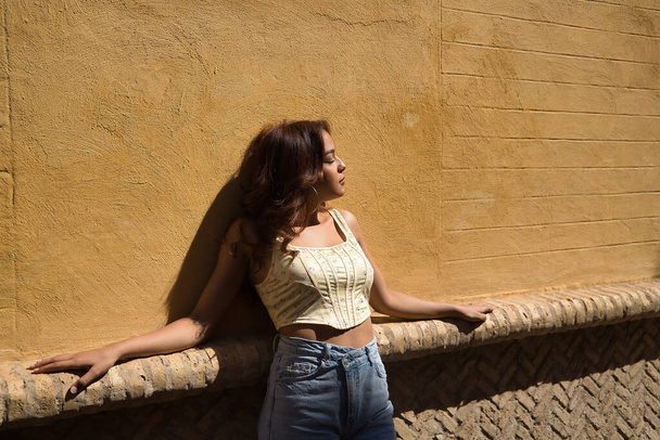 Young and beautiful woman with long brown hair and cinnamon skin leaning on a brick bench with a yellow wall in the background, with her eyes closed, relaxing in the sun. Concept beauty, tranquility. - Photo, Image