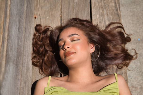 Portrait of beautiful young woman with long brown hair and cinnamon skin, lying on a wooden bench with her eyes closed. Beauty concept, makeup, hairstyles, fashion, trend. - Zdjęcie, obraz