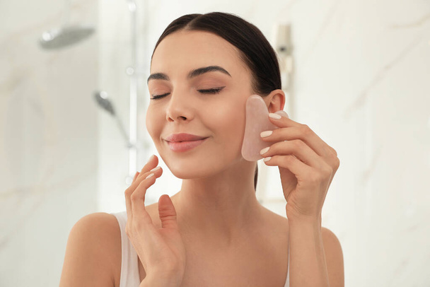 Schöne junge Frau tut Gesichtsmassage mit gua sha Werkzeug im Badezimmer - Foto, Bild
