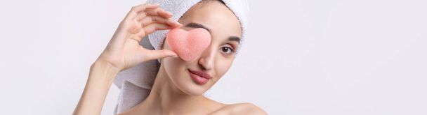 Beautiful girl with thick eyebrows and perfect skin at white background, towel on head, beauty photo. Holding a cosmetic pink heart sponge. - Photo, Image