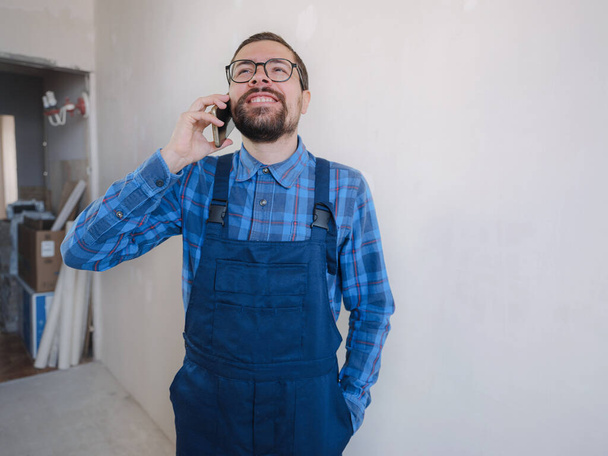 Egy kék munkaruhás fiatalember a javító lakásban. Lakásfelújítási koncepció. Férfi birtoklás video hívás okostelefon felett fehér beton fal háttér - Fotó, kép