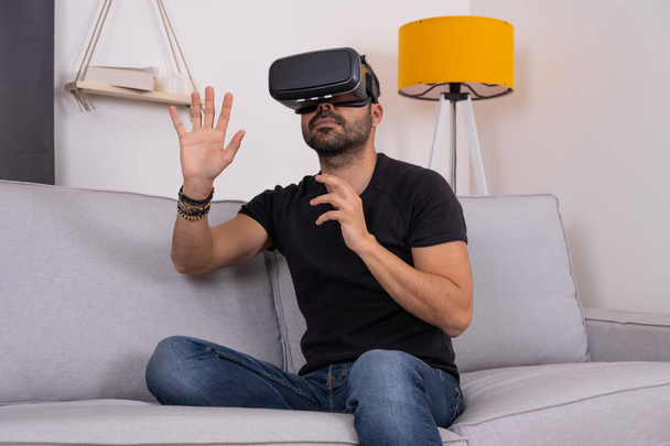 A fiatalember VR szemüveges fejhallgatót használ otthon, gesztikulálva. Virtuális valóság, jövőbeli technológia, oktatási videojáték. - Fotó, kép
