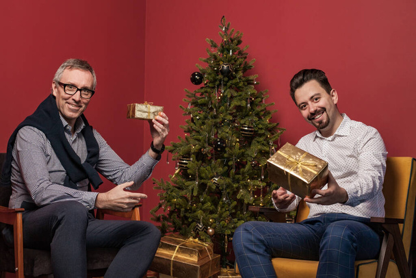 Привабливий літній чоловік і його син посміхаються і показують різдвяні подарунки на задньому плані червоної стіни і ялинці. - Фото, зображення