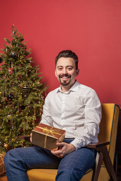 Mann im weißen Hemd hält Weihnachtsgeschenk goldene Schachtel auf rotem Hintergrund und Weihnachtsbaum - Foto, Bild