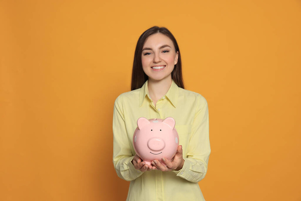 Happy young woman with piggy bank on orange background - Foto, imagen
