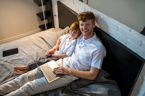 On internet. Ginger man and his cute son lying in bed with their gadgets - Valokuva, kuva
