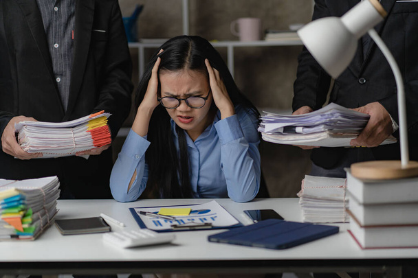 Aasialainen naispuolinen työntekijä työskentelee ylitöitä toimistossa myöhään yöllä Tyttö on ylityöllistetty ja hänellä on vaikeuksia stressin ja päänsäryn kanssa työskennellessään paperipinojen kanssa pöydällä.. - Valokuva, kuva