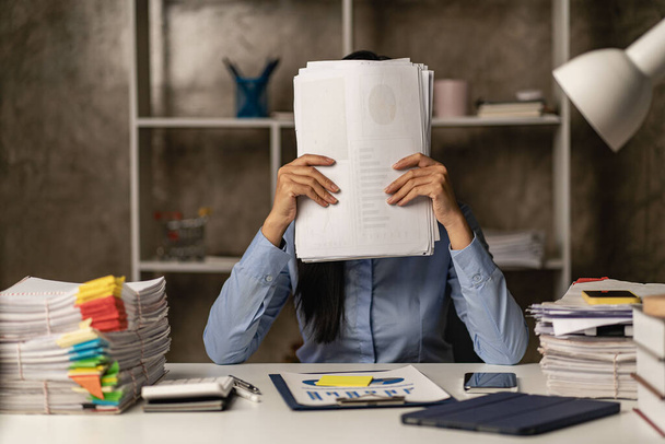 夜遅くにオフィスで残業しているアジアの女性労働者女の子は過労しており、机の上の書類の山で作業中にストレスや頭痛を感じるのに苦労しています. - 写真・画像