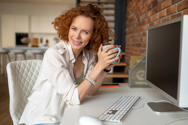 Op kantoor. Jonge vrouw die koffie drinkt in het kantoor terwijl ze achter de computer zit - Foto, afbeelding