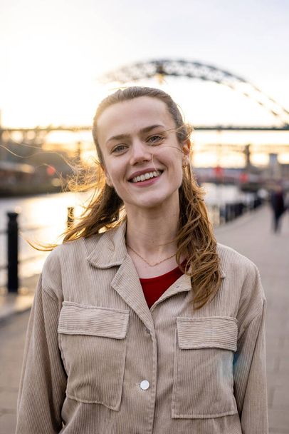 A portrait of a young woman spending the day in Newcastle Upon Tyne. She is looking at the camera and smilling while the sun shines behind her. - Foto, imagen