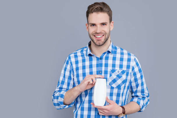 happy man with unbranded cosmetic in studio. unbranded cosmetic for man. stylish man hold bottle of unbranded cosmetic. man in checkered shirt hold unbranded cosmetics isolated on grey background. - Zdjęcie, obraz