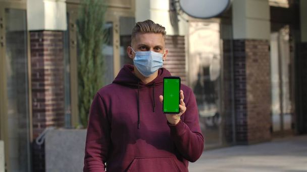 Portrait of young man in medical mask points to camera smartphone with green screen chroma key. Attractive blond man in a purple sweatshirt outdoors on the background of a blurred building. COVID-19 - 写真・画像