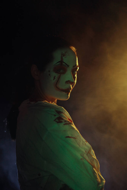 Junge Frau porträtiert blutrünstigen Zombie mit Horrorwunden im Gesicht und blutiger Kleidung vor dunklem Hintergrund mit Rauch. Gruseliges Bild zu Halloween. Porträt. - Foto, Bild