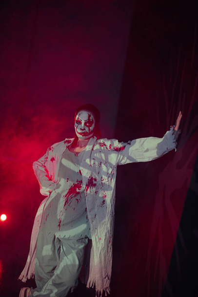 Jonge vrouw portretteert bloeddorstige zombie met horror wonden op haar gezicht en bebloede kleren tegen rode rook achtergrond. Enge afbeelding voor Halloween. - Foto, afbeelding