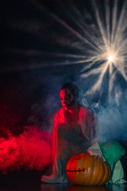 Jeune femme tient Jack o lanterne de citrouille et dépeint zombie sanguinaire avec des blessures horribles sur son visage sur fond sombre avec rétro-éclairage et fumée rouge. Image effrayante pour Halloween. - Photo, image