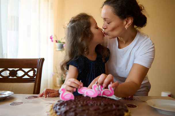 Любящая мать целует своего очаровательного прекрасного ребенка, дочь, поставив свечи на торт на день рождения. Именинница на своей пятой годовщине. Счастливого беззаботного детства. Счастливые семейные отношения - Фото, изображение
