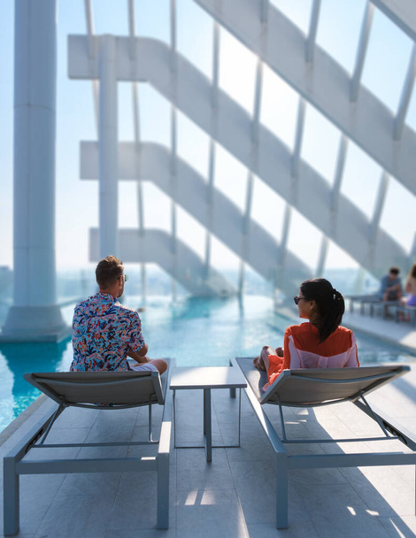 δύο άνδρες και γυναίκες χαλαρώνουν σε μια πισίνα στον τελευταίο όροφο ενός πολυτελούς ξενοδοχείου. Ασιατικές ταϊλανδέζες γυναίκες και καυκάσιοι άνδρες χαλαρώνουν δίπλα στην πισίνα σε μια ταράτσα - Φωτογραφία, εικόνα