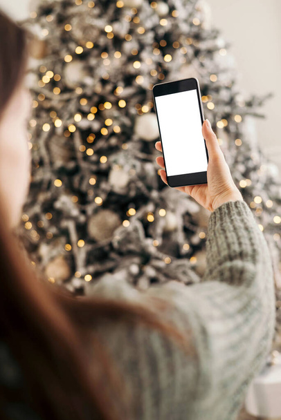 Young woman congratulating and holding phone and looking at camera background of Christmas tree at cozy living room with festive interior. mock-up. - Zdjęcie, obraz