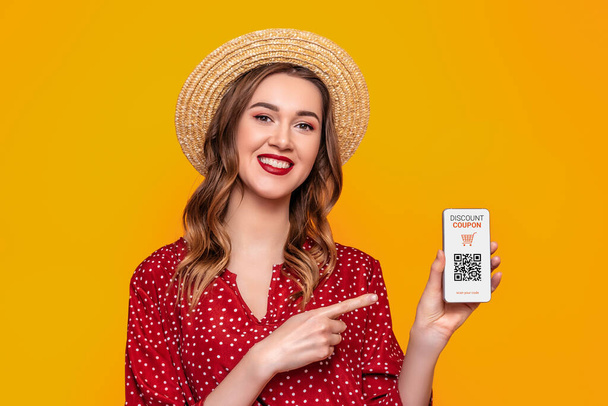 portret młodej białej kobiety trzymającej telefon komórkowy z kuponem rabatowym i kodem QR odizolowanym na pomarańczowym tle w studio.. - Zdjęcie, obraz