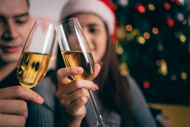 Mutlu Asyalı çift şampanya kızartıyorlar, akşamları koltukta oturuyorlar ve arka planda bir Noel ağacı ve ışıklar var. Noel arifesinde sevgili - Fotoğraf, Görsel