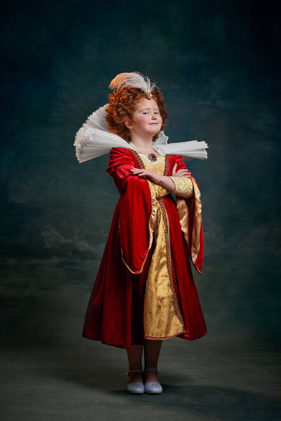 Portrait of little red-headed child in baroque costume of royal person isolated over dark green background. Pride. Concept of historical remake, comparison of eras, medieval fashion, emotions, queen - Photo, image
