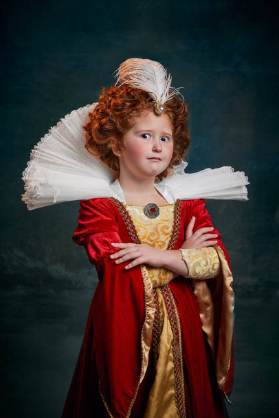 Portrait of little red-headed girl, child in costume of royal person isolated over dark green background. Shocked emotions. Concept of historical remake, comparison of eras, medieval fashion, queen - Foto, afbeelding