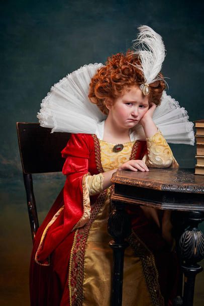 Portrait of little red-headed girl in costume of royal person sitting in tears isolated over dark green background. Concept of historical remake, comparison of eras, medieval fashion, emotions, queen - Fotoğraf, Görsel