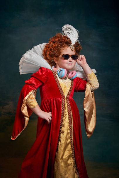 Portrait of little red-headed girl, royal person in sunglasses and headphones isolated over dark green background. Concept of historical remake, comparison of eras, medieval fashion, emotions, queen - Photo, image