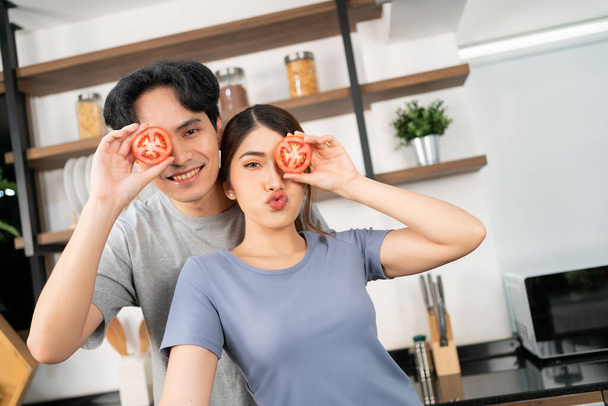 Healthy young Asian couple holding a fresh sliced tomato in their eye with a smile and happy emotion. An active lifestyle with healthy and vegetarian food advertisement. Image with copy space. - Photo, Image