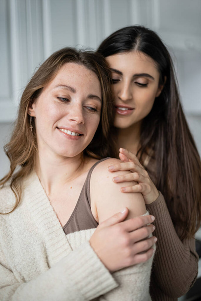 Усміхнена лесбіянка обіймає партнера в теплому светрі вдома
  - Фото, зображення