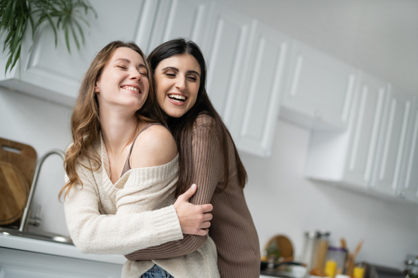 Cheerful lesbian woman hugging young girlfriend in kitchen  - Zdjęcie, obraz