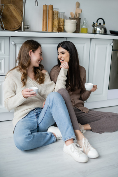 Молодые лесбиянки держат чашки кофе, разговаривая на полу на кухне  - Фото, изображение