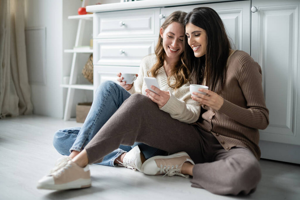 Lesbian women holding cups of coffee and using smartphone on floor in kitchen  - Foto, Bild