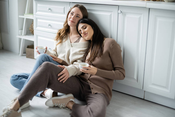 Lesbian women holding cups of coffee while sitting on floor in kitchen  - Photo, Image