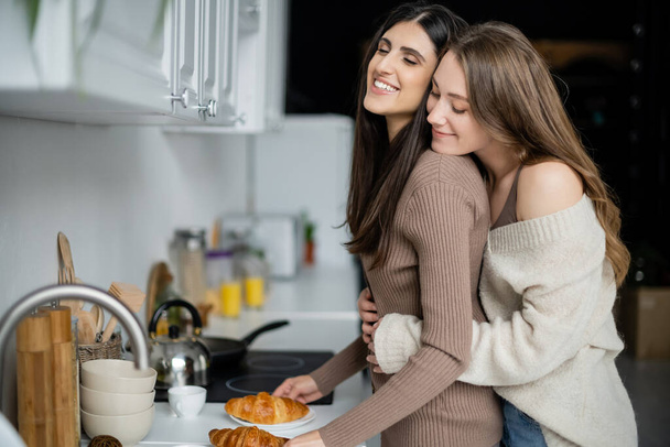 Positive lesbian woman in cozy sweater hugging girlfriend near croissants in kitchen - Photo, Image