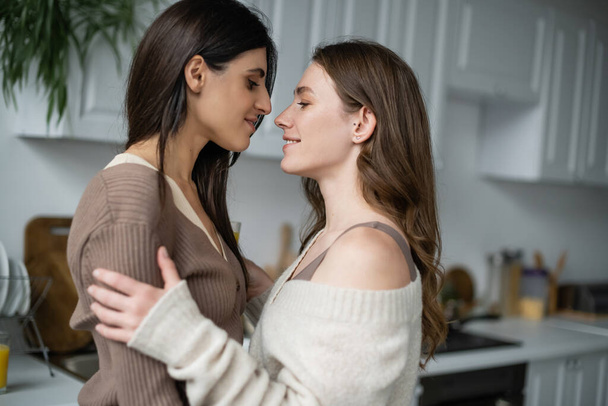 zijaanzicht van lesbisch paar knuffelen en kijken naar elkaar in keuken  - Foto, afbeelding