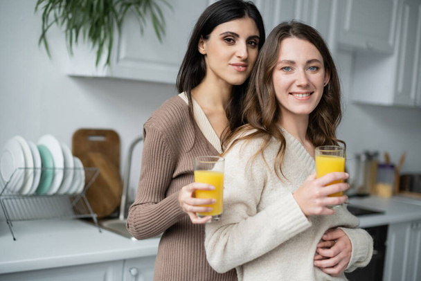 Χαμογελαστό λεσβιακό ζευγάρι που κρατάει ποτήρια χυμό πορτοκαλιού στην κουζίνα  - Φωτογραφία, εικόνα
