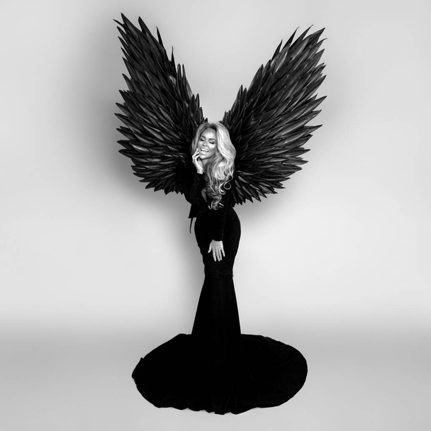 エレガントなロングブラックのドレスと大きな翼の見事なブロンドの女性がスタジオでポーズをとっています。豪華な夜のファッション。グラマーファッションモデル。翼を持つ女性。黒天使。白黒写真. - 写真・画像
