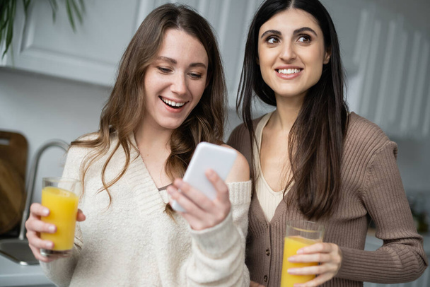 Smiling woman using smartphone and holding orange juice near girlfriend in kitchen  - Foto, Imagen