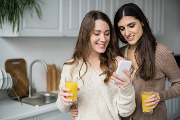 Same sex couple holding orange juice and using smartphone in kitchen  - Foto, Bild